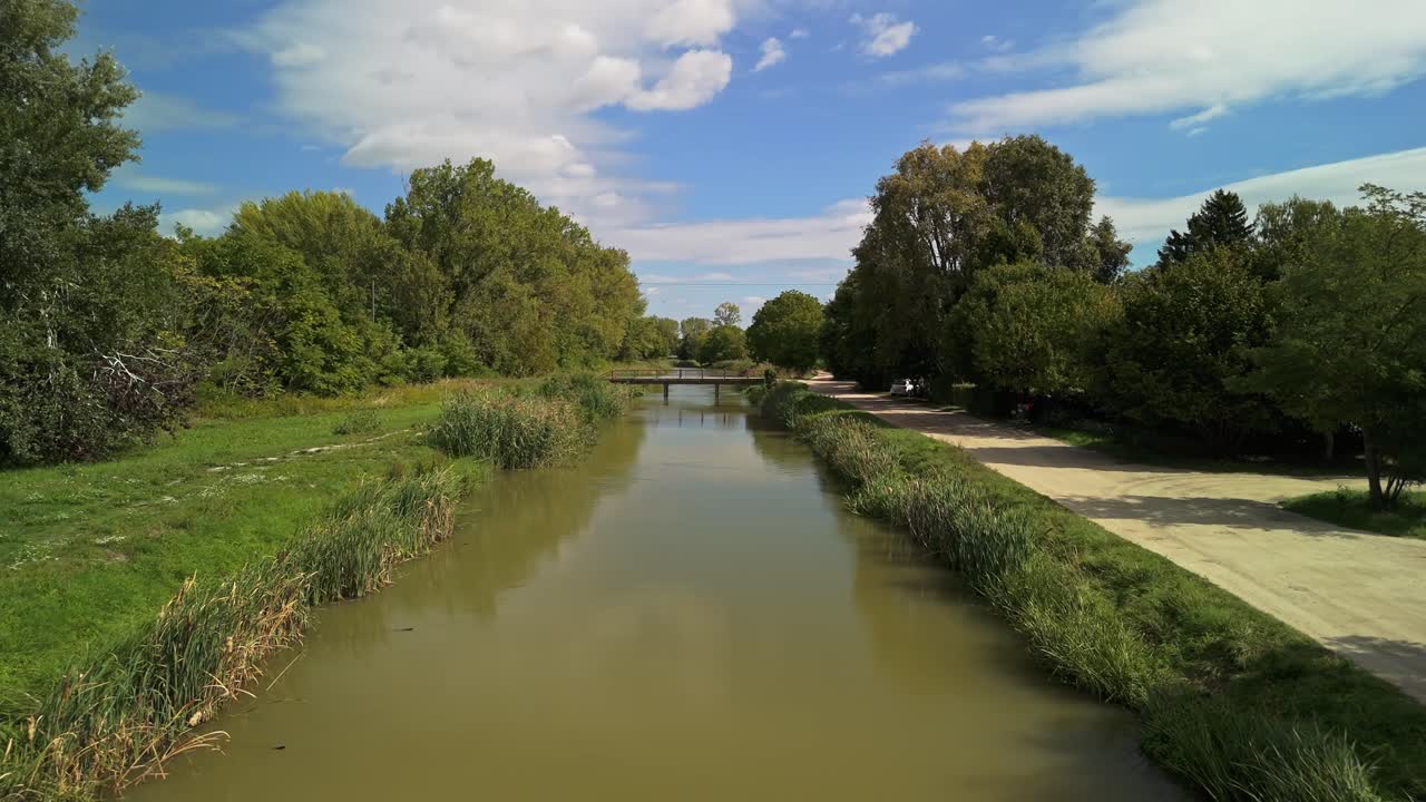 Peaceful aerial view of the small belt canal in Dömsöd, Hungary, lined with lush green trees and a quiet riverside dirt road under bright early autumn skies