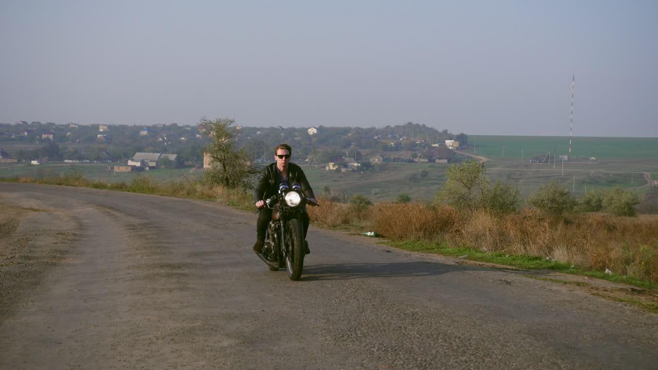 vista frontal de un joven elegante y fresco con gafas de sol y chaqueta de cuero conduciendo su helicóptero en una carretera de asfalto en un día soleado