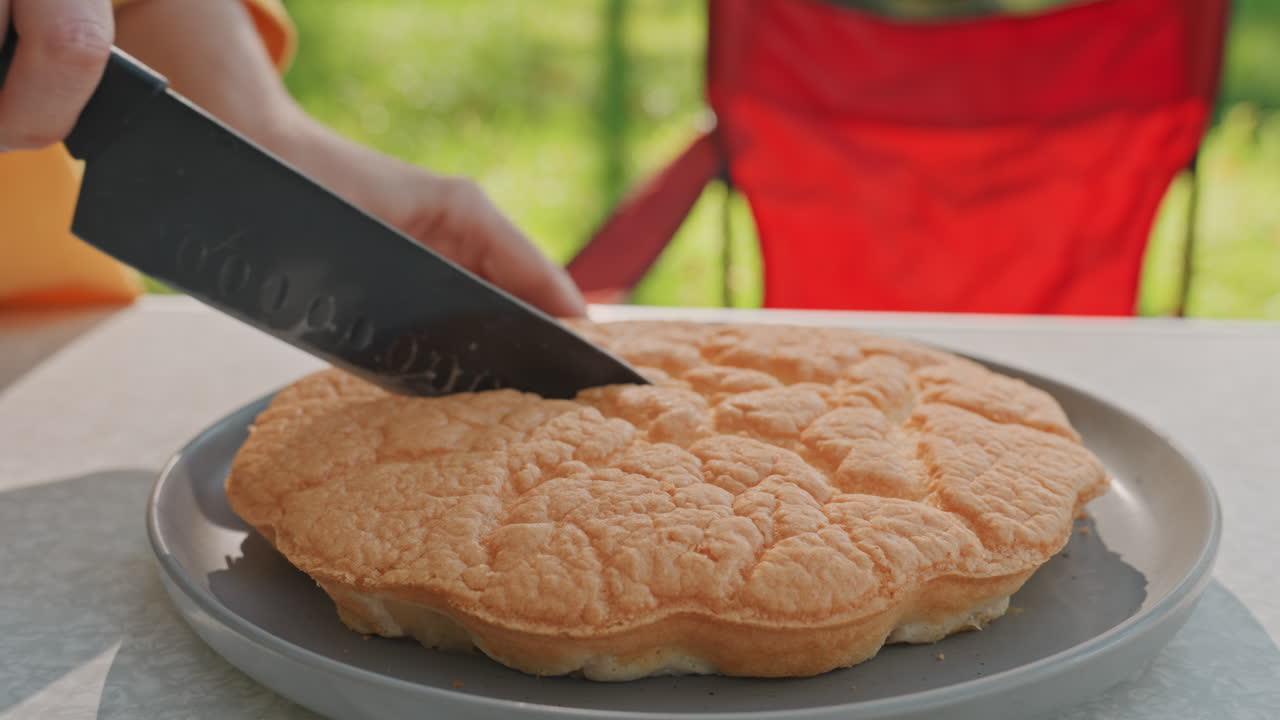 Outdoor camping table with bread being sliced by camper chef closeup serrated knife cutting soft cloud bread on plate red camp chair and green trees in background gentle morning light and simple