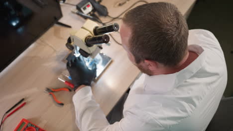 una vista hacia abajo de un técnico con una bata blanca de laboratorio y guantes negros trabajando en un circuito bajo un microscopio en un escritorio en un laboratorio, visto desde arriba