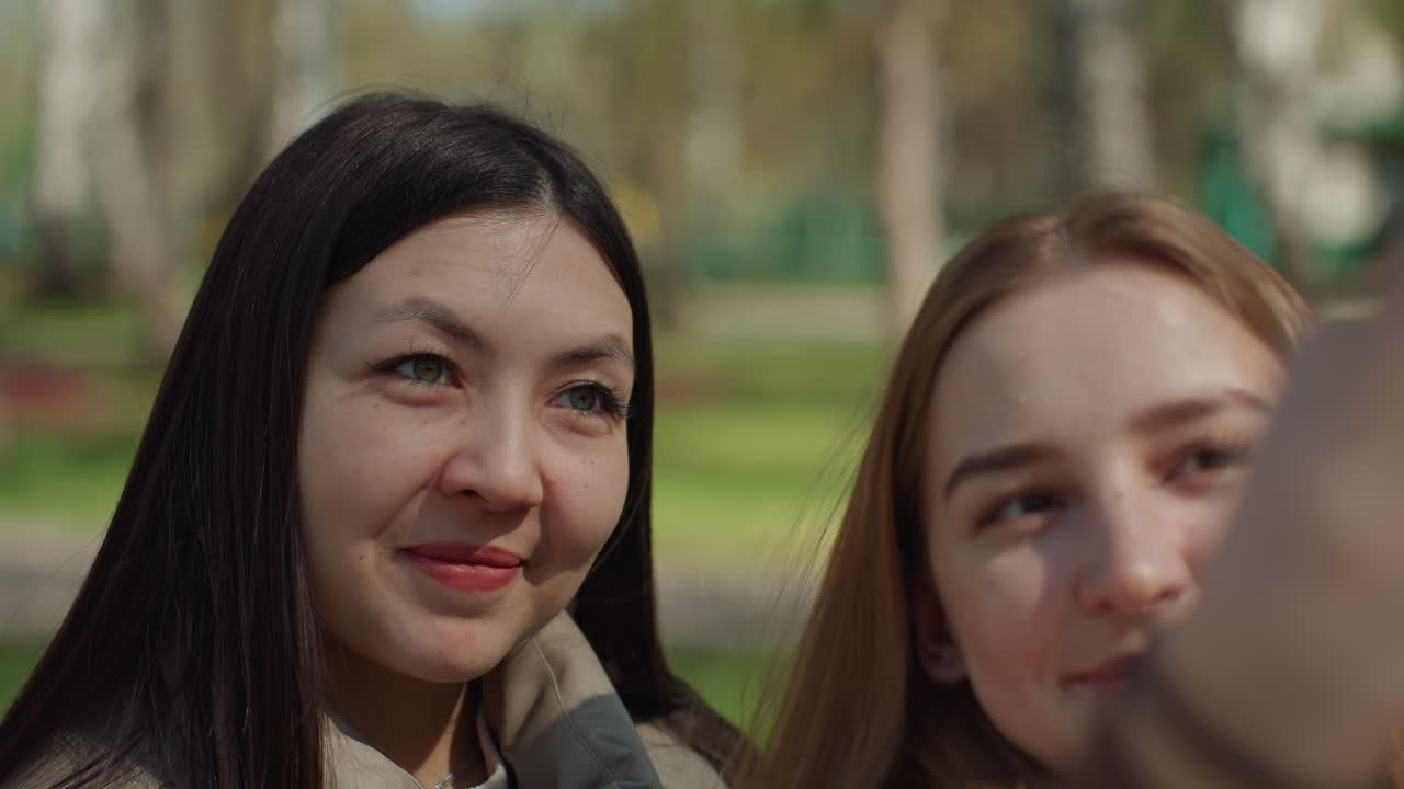 아시아 여성과 백인 친구가 공원 벤치에서 셀카를 찍기 위해 포즈를 취하고 있고, 스마트폰 프레임에는 웃는 얼굴이 담겨 있으며, 장난기 넘치는 표정과 햇빛, 나무, 캐주얼한 패션 스타일을 보여주는 인플루언서의 분위기가 느껴져요.