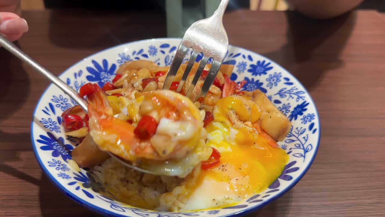 Person lifting shrimp, egg, and rice from colorful bowl with utensils on wooden table indoors