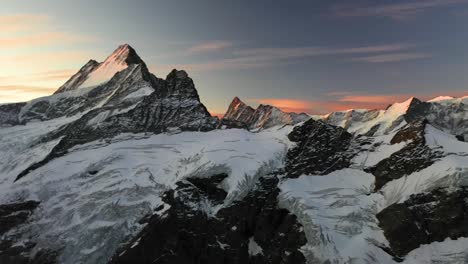 Drone-shot-panning-from-right-to-left-over-the-snow-covered-mountain-peaks-in-the-alps-of-Switzerland-during-a-colorful-and-dramatic-sunrise-with-red-and-orange-filled-sky