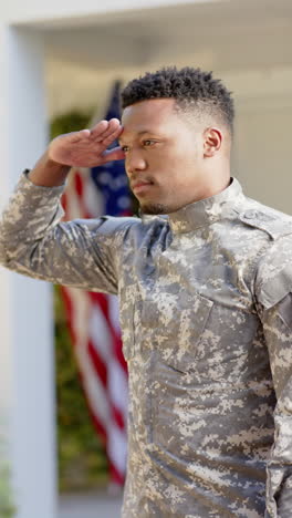 vertical video of happy african american male soldier saluting at home, slow motion