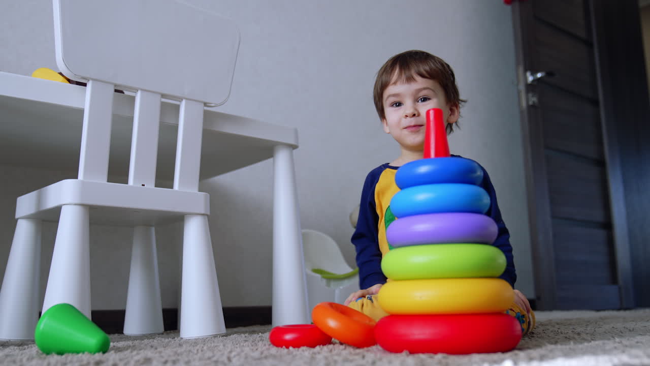 Beautiful smiling toddler boy assembling colorful pyramid at home. Cute child turns over the toy. Low angle view.