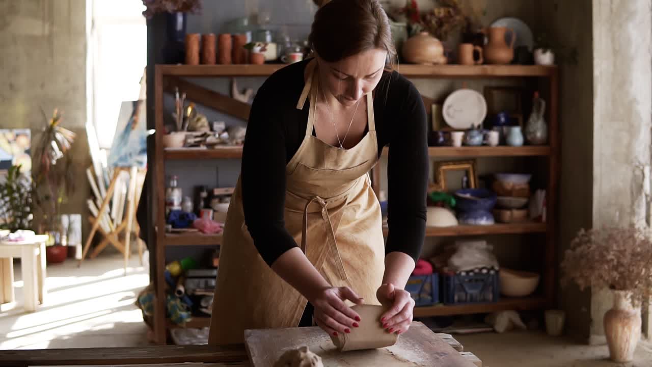 Front view of female potter wearing beige apron kneading softly clay piece on worktop, working with her hands. Pottery products on shelves behind, lens flares