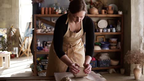 front view of female potter wearing beige apron kneading softly clay piece on worktop, working with her hands. pottery products on shelves behind, lens flares