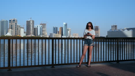 Beautiful-girl-with-long-hair-in-sunglasses-using-smartphone-app-at-sunset-river-quay-near.