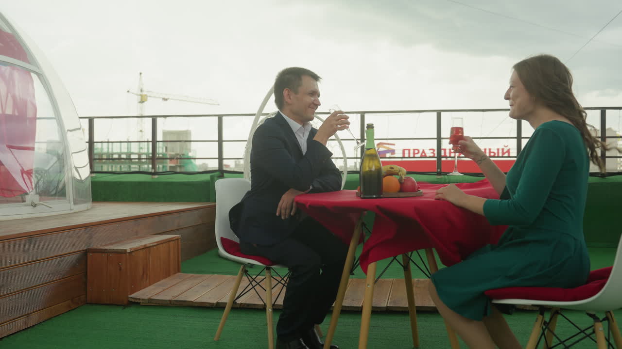 Cena intima su un tetto con cupola trasparente. Coppia bianca di dirigenti e esperti di marketing che brinda con il vino, tovaglia rossa, bottiglia e bicchieri, skyline della città sullo sfondo, luce di candela sul prato verde, riservato