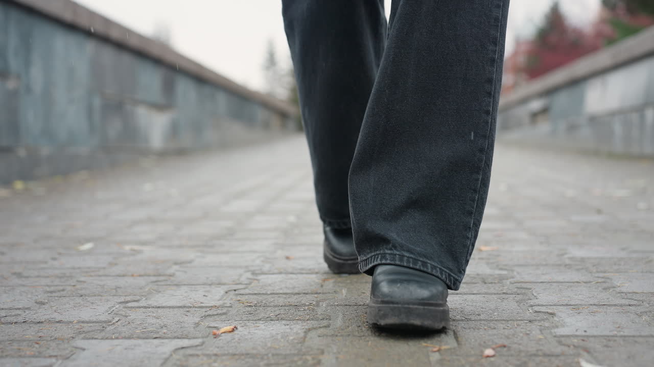 Close up of someone's black trousers and black boots walking slowly toward camera on wet paved urban path, face not visible, capturing motion of steps and rainy atmosphere in quiet autumn setting