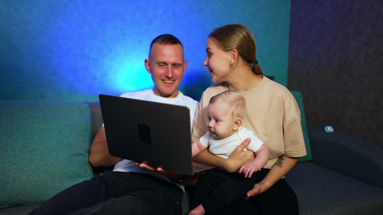 Modern Caucasian couple with little baby sitting in front of laptop. Cute infant boy looks at computer screen with surprise.