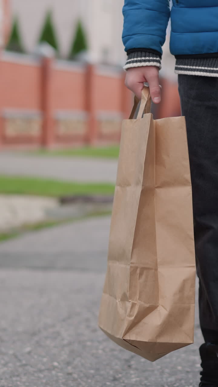 vista de ángulo inferior de un individuo con chaqueta azul y pantalones vaqueros caminando en un área residencial, llevando una bolsa de papel, árboles de otoño, casas, en el fondo con vegetación