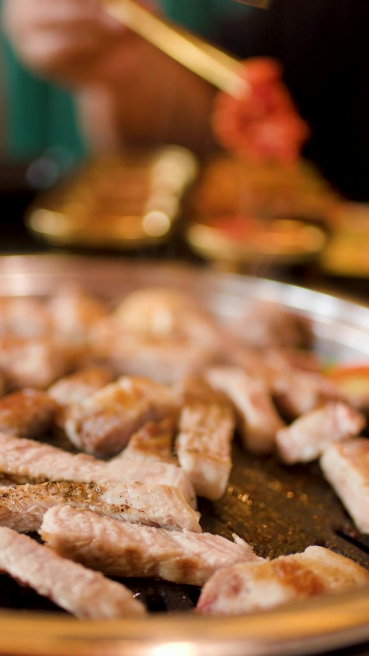 Close-up daging babi yang dipanggang di atas hotplate, tangan menggunakan penjepit, pencahayaan hangat, kedalaman bidang dangkal