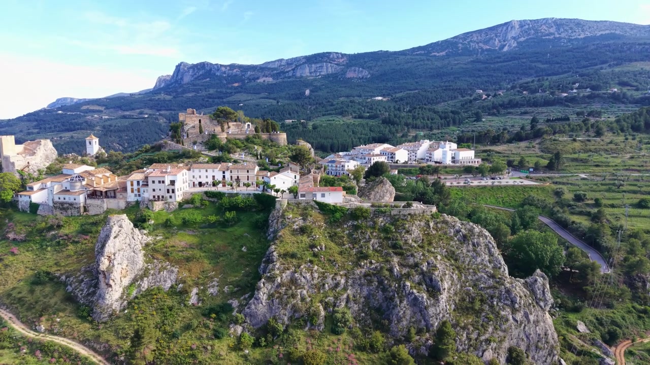 Villaggio-castello di Guadalest arroccato su una collina rocciosa ad Alicante, Spagna, circondato da montagne verdi, edifici bianchi e rovine medievali visibili dall'alto.