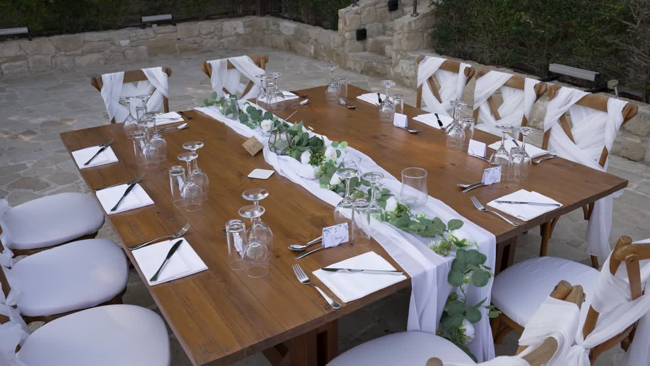 An inviting outdoor table is elegantly set up with white decorations in a lush garden. The wooden table features stylish chairs adorned with flowing fabric, creating a perfect atmosphere