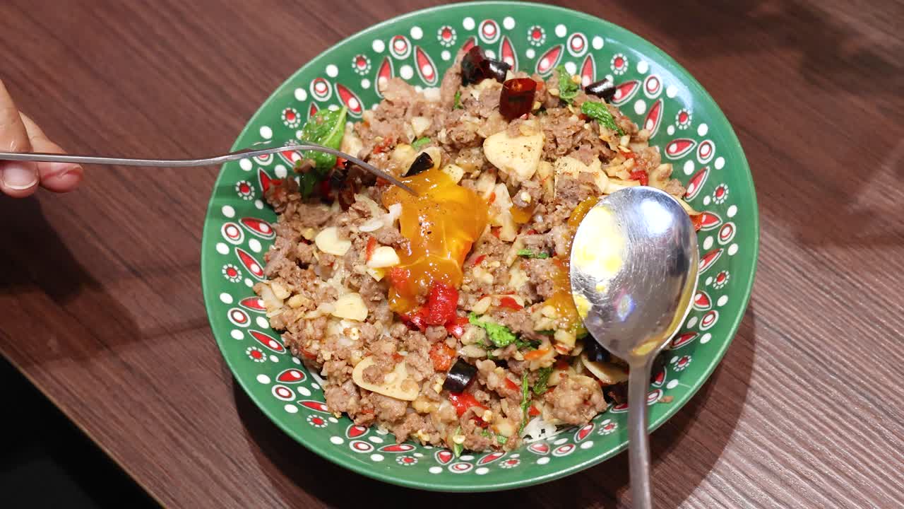 Hands use a fork and spoon to mix a colorful Thai rice dish with egg, beef, garlic, chili, and herbs under bright indoor lighting