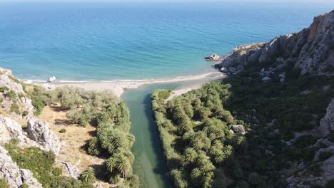 Aerial-view-of-emerald-river-across-the-rocky-mountains-with-palm-tree-vegetation-with-Mediterranean-sea-on-the-background