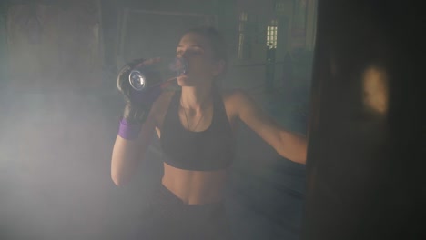 Thirsty-female-boxer-taking-a-break-drinking-from-the-water-bottle-after-training-in-the-dark-gym-with-smoke.-Beautiful-woman