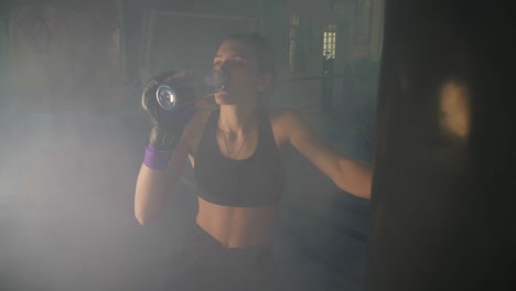 boxeadora sedienta tomando un descanso bebiendo de la botella de agua después de entrenar en el gimnasio oscuro con humo. mujer hermosa