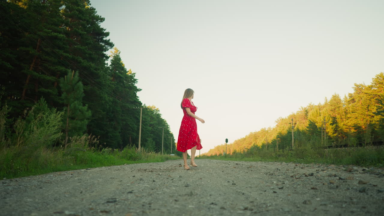 Woman in red dress walking along rural dirt road turning head to look behind, surrounded by tall green trees and utility poles, with sunlight casting soft glow in tranquil countryside landscape