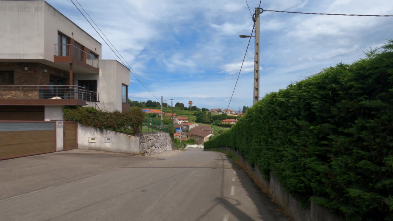 Quiet residential street in Gijon countryside. Modern house, hedge, concrete road, and hillside town visible under blue sky.