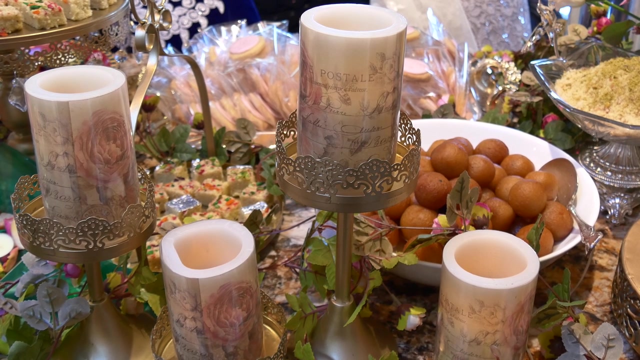 Ornate dessert spread featuring Gulab Jamun, pistachio-topped pastries, and wrapped treats, beautifully accented with vintage-style pillar candles on gold stands and lush greenery.