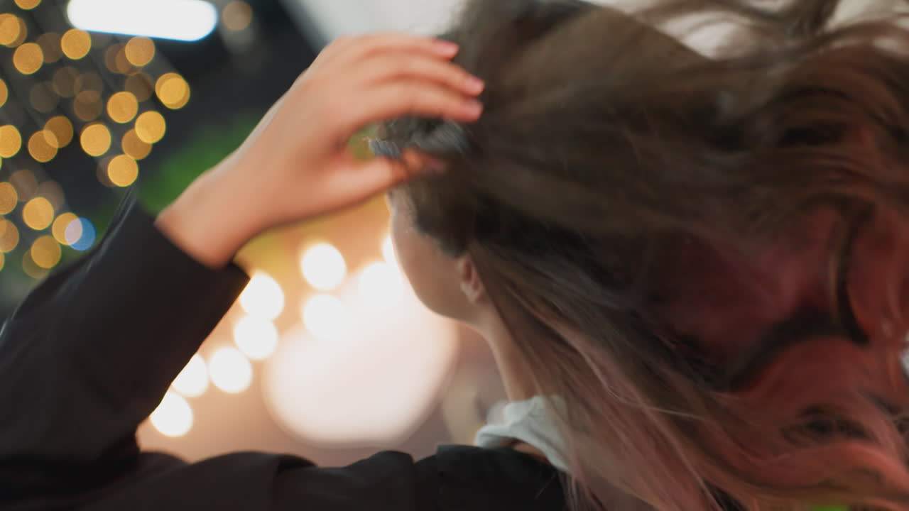 Mladá studentka si svazuje culík za bokeh světel, brunetka s růžovými proužky si upravuje vlasy v útulné vnitřní scéně, ruce upravují dlouhé kadeře, teplá jemná záře a reflexivní nálada, okno koleje
