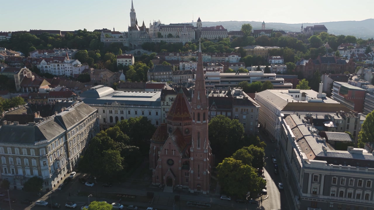 Aerial circling Budapest’s Szilágyi Dezső Square Reformed Church, showcasing its tall spire and distinctive red-tiled roof with the Fisherman’s Bastion unfolding in the background.
