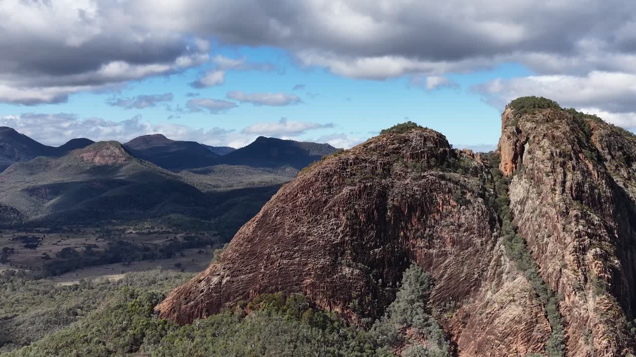 Drone camera smoothly pans across Split Rock, revealing rugged cliffs, forested valleys, and distant mountain ranges under partly cloudy daylight skies