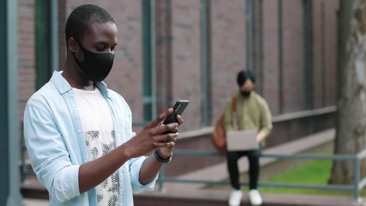 African american student wearing facial mask and using his smartphone in the street near the college