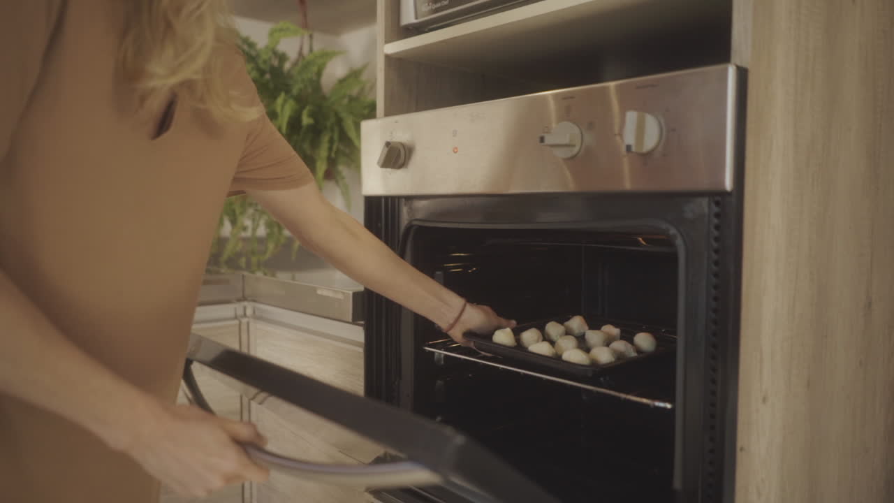Person baking traditional chipa bread at home, placing a tray of dough balls into the oven in a warm, inviting kitchen environment