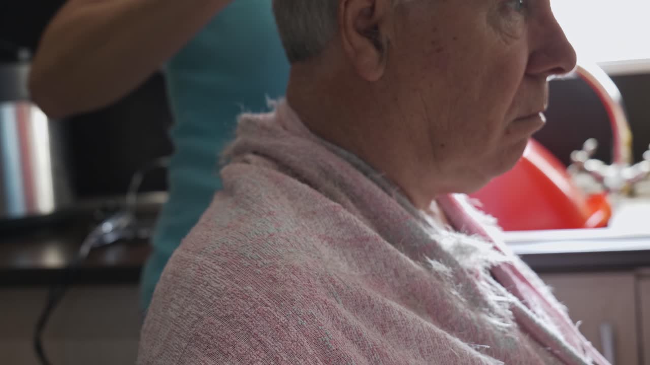 Head profile of grey haired old man having haircut in home kitchen by amateur barber, left to right slide close up.
