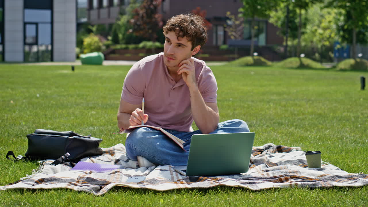 Focused man studying campus lawn with laptop. Thoughtful student writing notes