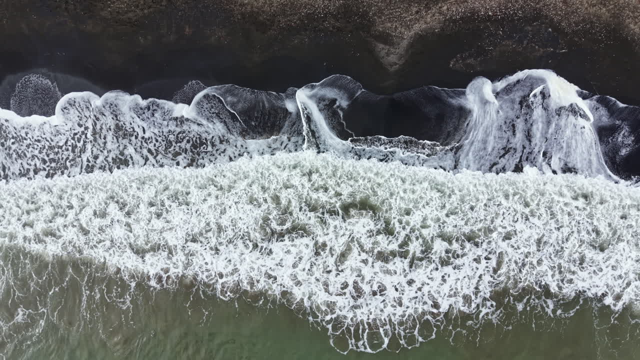 Onde che si infrangono su una spiaggia di sabbia nera con schiuma bianca e limpida sulla battigia, di giorno, in una zona costiera.