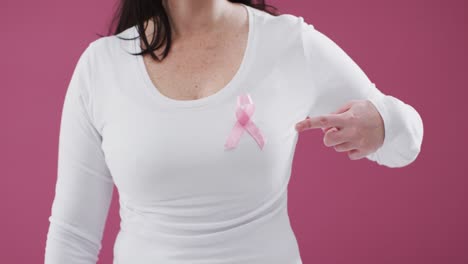 mid section of a woman pointing to the pink ribbon on her chest against pink background