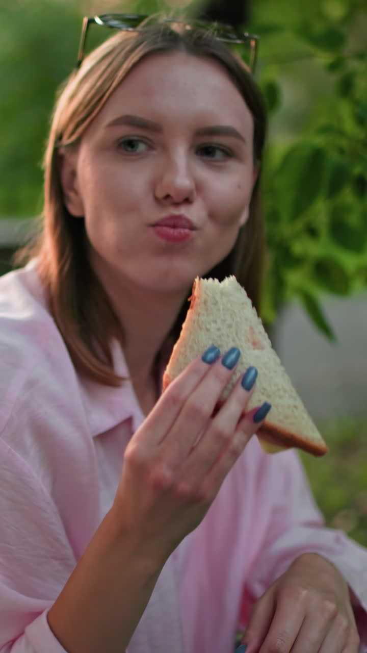 primer plano de una mujer disfrutando de un sándwich con una sonrisa en la cara, rodeada de naturaleza verde en un entorno al aire libre relajado, con una vista parcial de algunos sándwiches en la mano