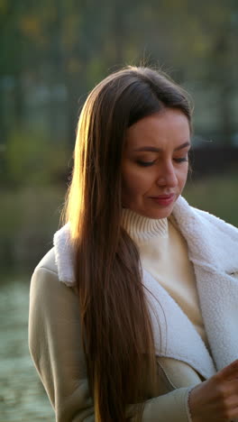 relaxed woman leaned on the handrail of the bridge. brunette lady using her phone near the river in autumn. blurred backdrop. vertical video