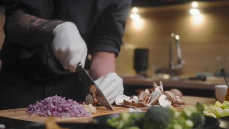 mushrooms being chopped on a wooden board by a young male chef in an elegant black shirt with tattoos wearing white gloves