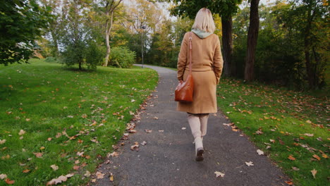 Una-Mujer-Con-Un-Abrigo-Y-Una-Bolsa-Camina-En-Un-Parque-De-Otoño,-Vista-Trasera