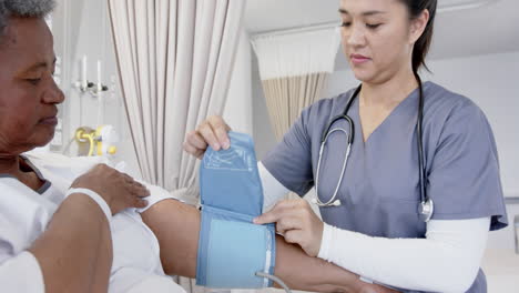 diverse female doctor testing blood pressure of senior female patient in hospital room, slow motion