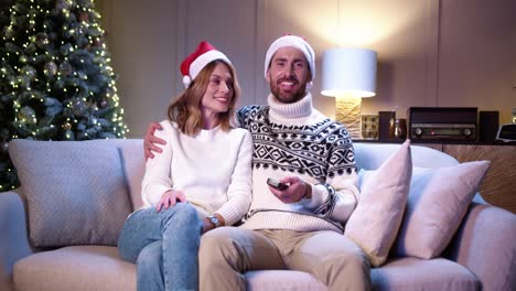 familia feliz pareja casada con sombreros de santa sentados juntos en una casa acogedora cerca de un árbol de navidad decorado y viendo televisión en año nuevo