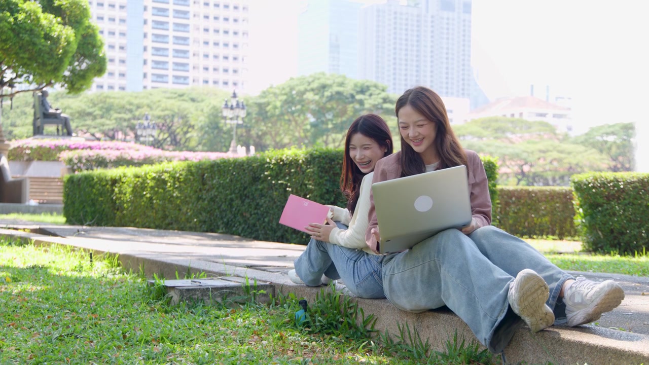 明るい日差しの下で、2人の若い女性が屋外で背中合わせに座り、ラップトップとノートブックを使用している。