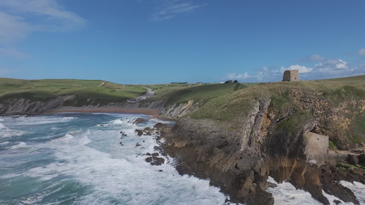 Drone flight over the 16th-century Santa Justa Hermitage and San Telmo Tower, built on Cantabrian cliffs near Ubiarco, surrounded by green meadows and the turquoise sea under soft coastal light