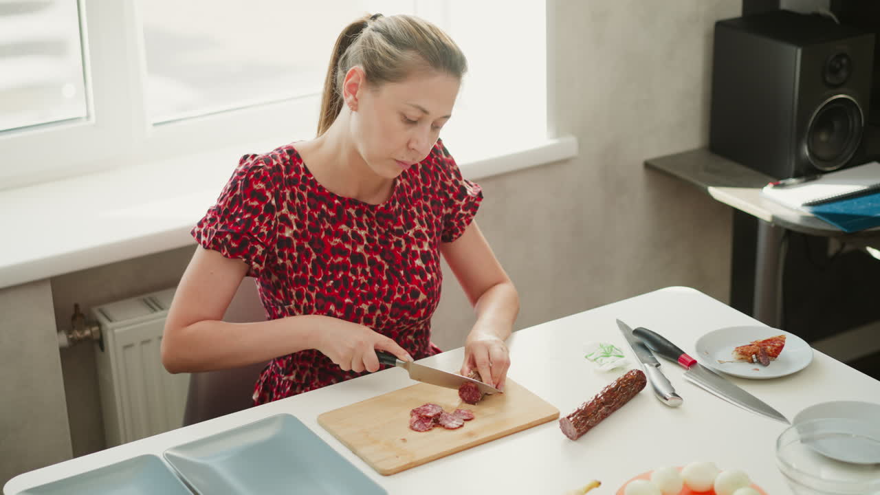 High angle of lady in red dress seated by bright window light, slicing dry cured meat into thin round pieces on wooden board over white table, showcasing precise knife technique and rich texture