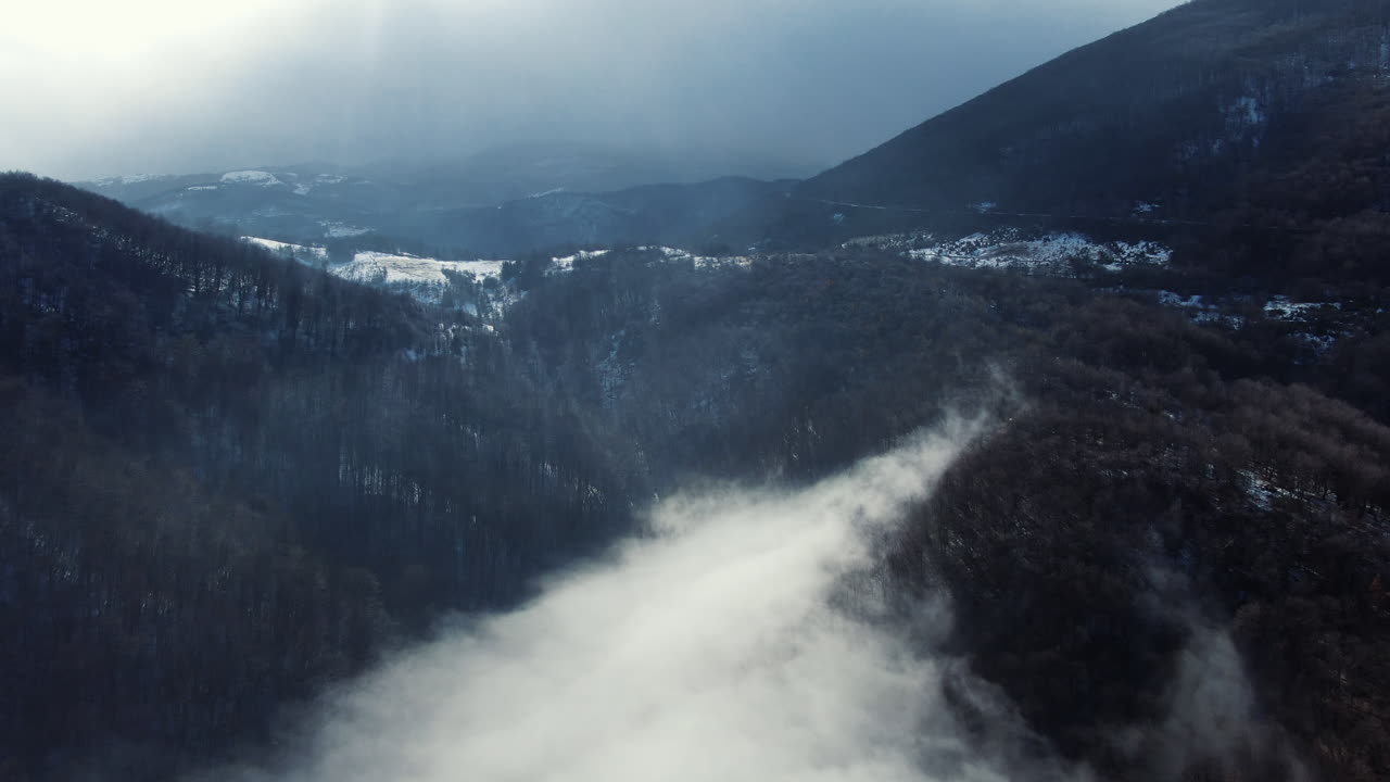 Mountains covered with trees appear through a blanket of clouds Some snow is visible on the peaks with a light sky behind the mountains