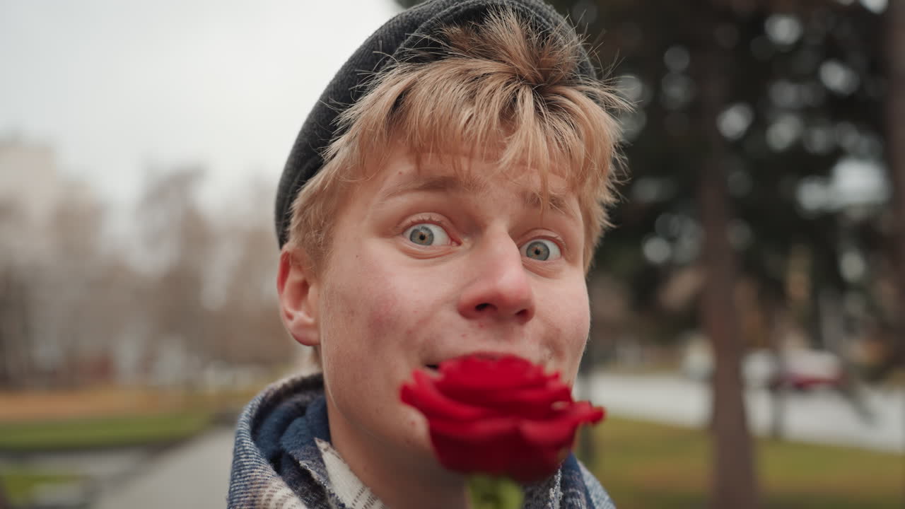 Young white creator comedian with rose in outdoor park, beanie and scarf, goofy grin and smirk, exaggerated expressions for comic effect, closeup shallow depth, lively energy and spontaneous charm