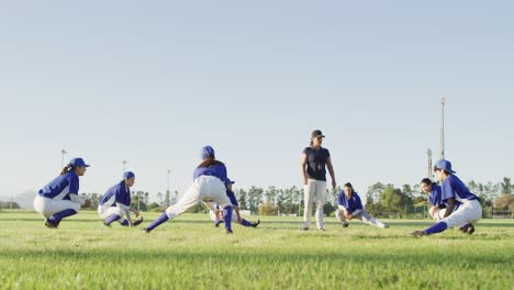 diverse group of female baseball players and coach warming up on pitch, squatting, stretching legs