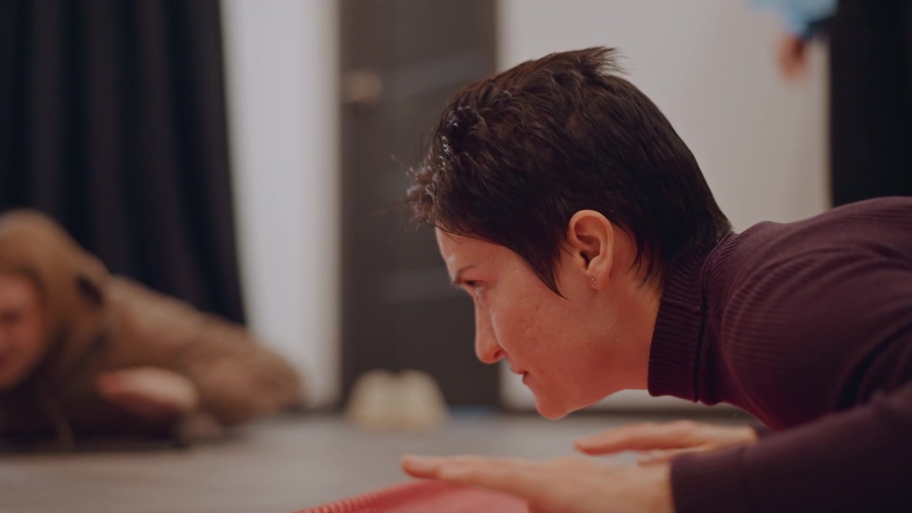 Donna asiatica su tappetino che pratica la respirazione. Lezione intima in studio con studentessa di musica dai capelli corti che estende i palmi, maglione bordeaux, espressione concentrata, istruttore sullo sfondo, espirazione lenta e controllata.