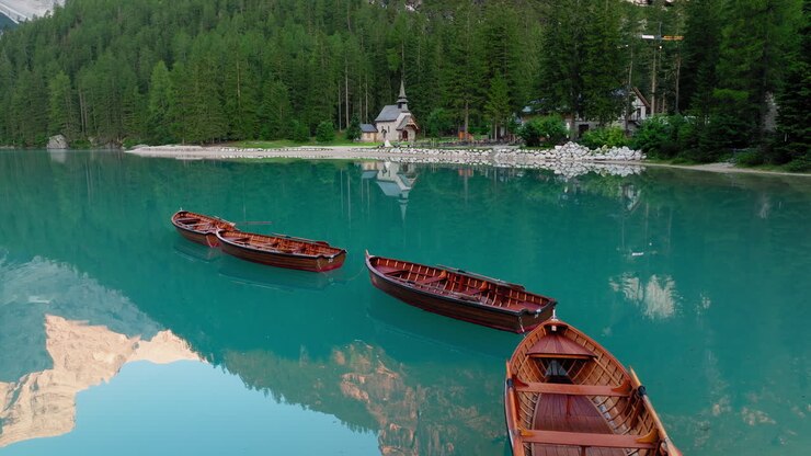 Slow panning drone shot of Lago di Braies at sunrise on a clear morning, with mirror like water reflecting the surrounding Dolomites and wooden boats moored beside the iconic boathouse.