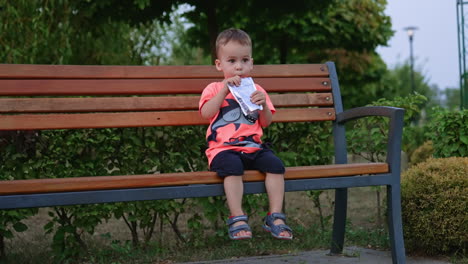 adorable caucasian baby sitting on the bench drinks juice. kid gives an empty pack to his mom.